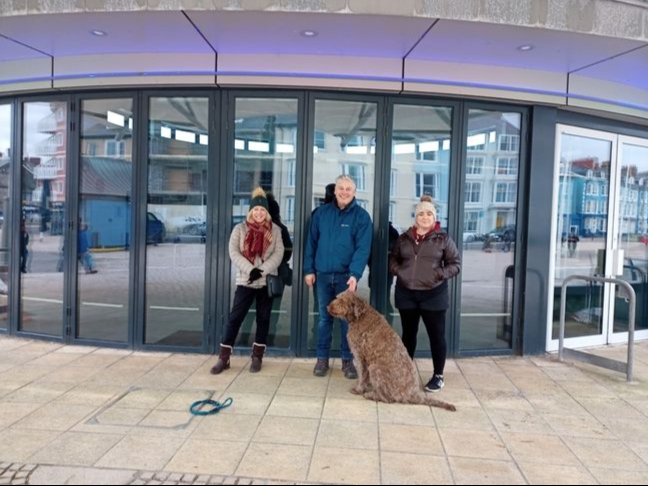 Caru Aber members Christine Copeland, Jeff Jones, and Bethan Thomas standing outside Aberystwyth bandstand, where the CANU ABER gyda CARU ABER music event will take place.